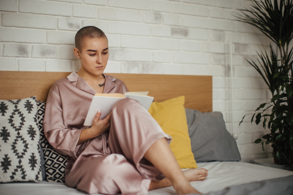 Young woman with cancer reading book. Concept of cancer awareness. Female cancer patient lying in bed, relaxing at home.