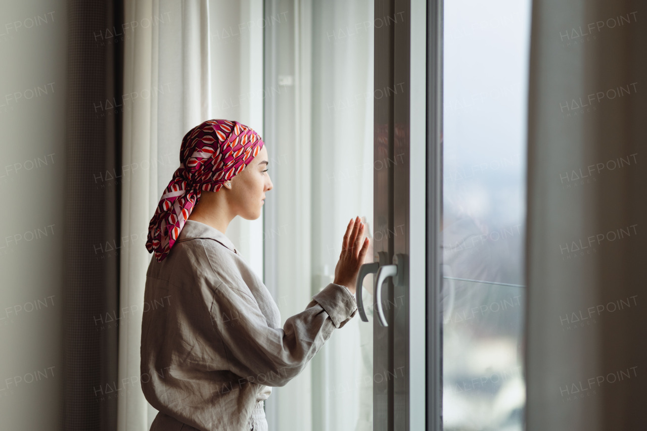 Young unhappy woman with cancer standing in front of the window.