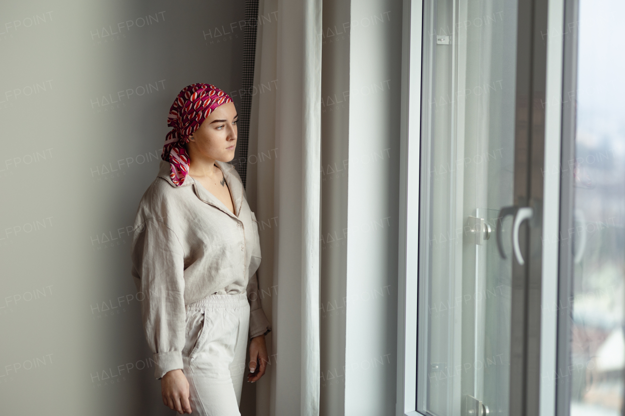 Young unhappy woman with cancer standing near the window and looking at view.