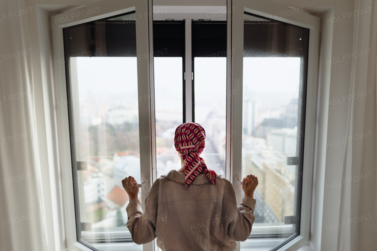 Rear view of young unhappy woman with cancer standing in front of the window.