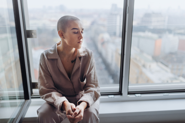 High angle view of young unhappy woman with cancer sitting in window.