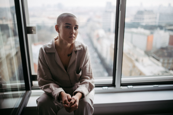 Young unhappy woman with cancer sitting in window and looking at view.