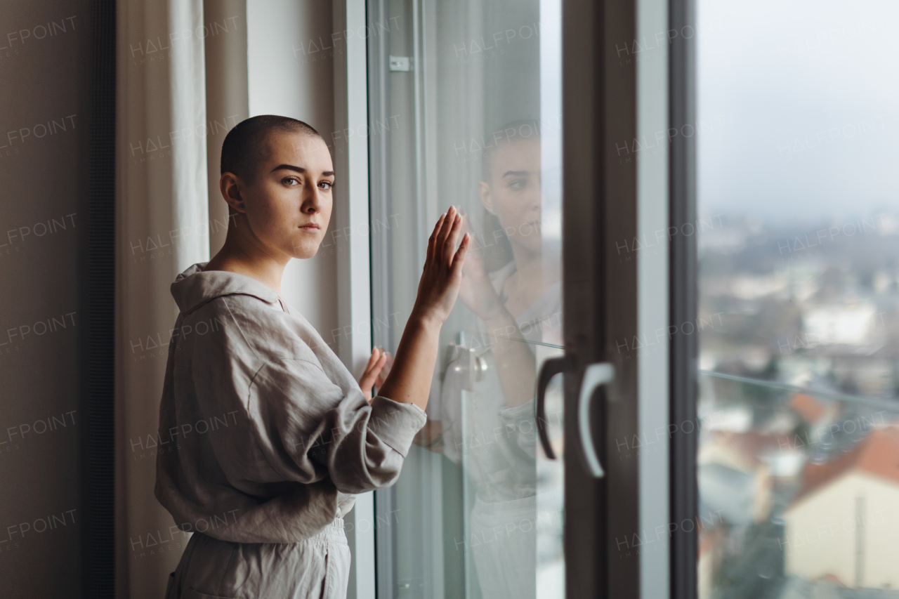 Young unhappy woman with cancer standing near the window and looking at view.