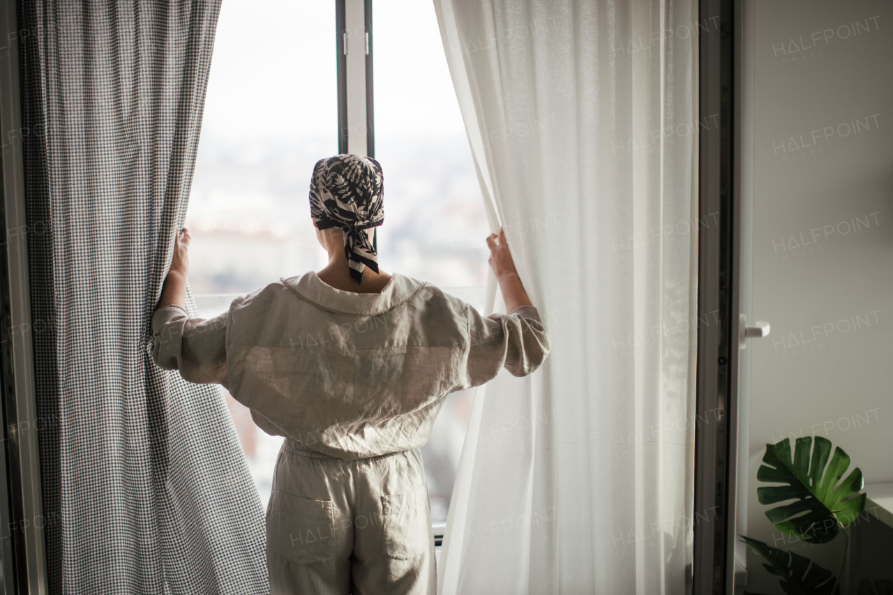 Rear view of young unhappy woman with cancer standing in front of the window.