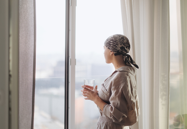 Young unhappy woman with cancer standing near the window and looking at view.