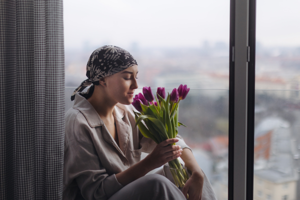 Young woman with cancer sitting in a window and smelling bouquet of tulips.