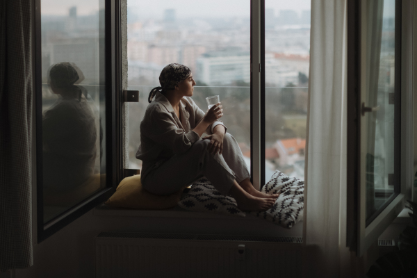 Young unhappy woman with cancer sitting in window and looking at view.