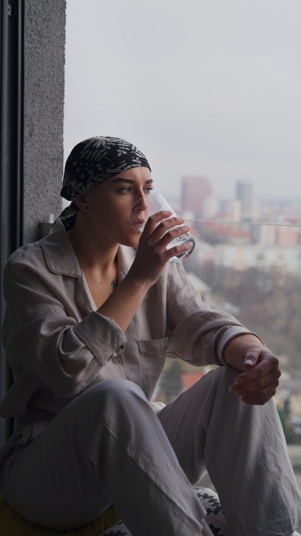 Young unhappy woman with cancer sitting in window, drinking water. Vertical footage.