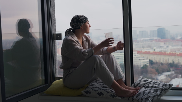 Young unhappy woman with cancer sitting in window, drinking water.