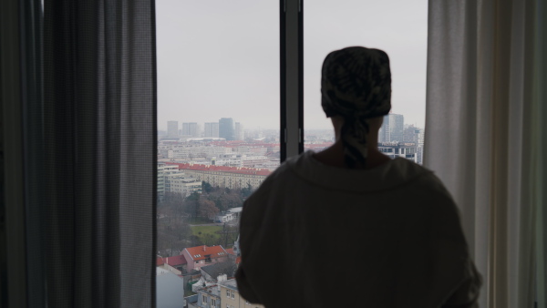 Young unhappy woman with cancer standing near the window and looking at view.