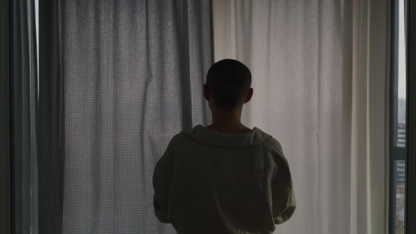 Young unhappy woman with cancer standing near the window and looking at view.