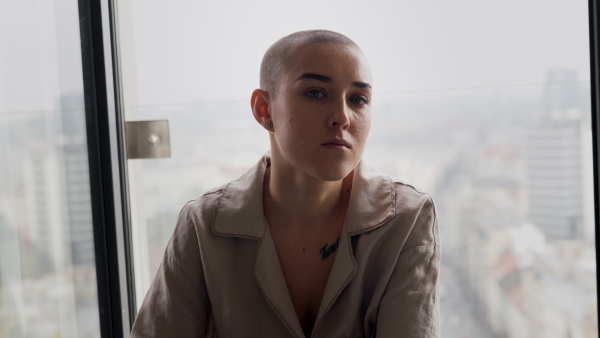 A young unhappy woman with cancer sitting near a window and looking at camera.