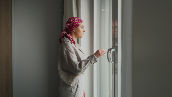 Young unhappy woman with cancer standing near the window and looking at view.