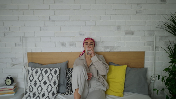 A young woman with cancer drinking water and lying in a bed, cancer awareness concept.