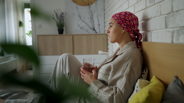 A side view of young woman with cancer drinking water and lying in a bed, cancer awareness concept.