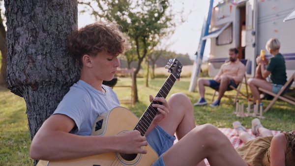 Family traveling in caravan, camping and exploring new places. Boy to playing guitar, sitting in front of a caravan.