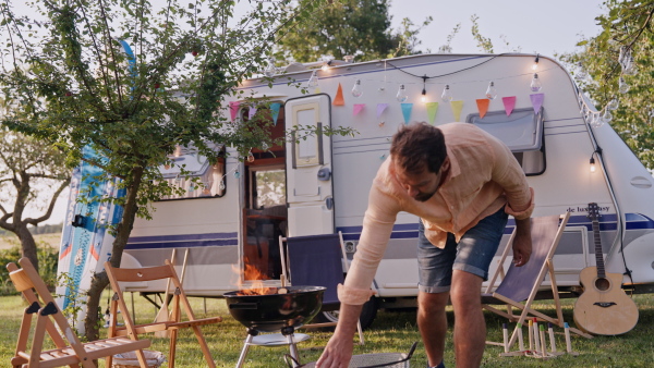 Dad making fire in fire pit, grill in front of a caravan, preparing diner. Family traveling in caravan, exploring new places.