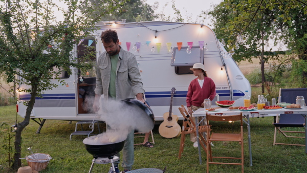 Dad grilling in front of a caravan, preparing diner at family camping trip in nature. Man holding gill bbq tongs, flipping sausages.