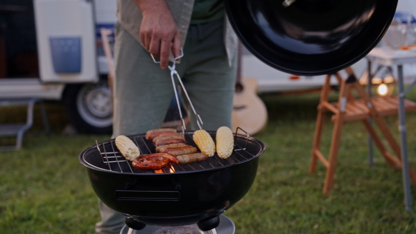 Dad grilling in front of a caravan, preparing diner at family camping trip in nature. Man holding gill bbq tongs, flipping sausages.