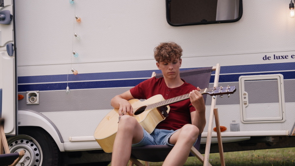 Family traveling in caravan, camping and exploring new places. Boy to playing guitar, sitting in front of a caravan.