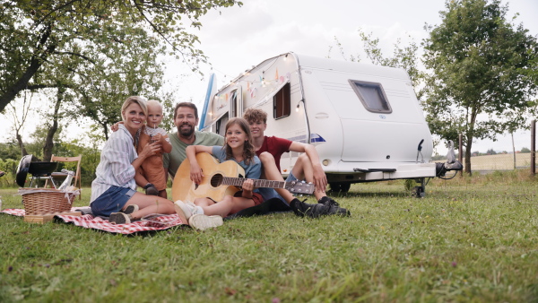 Family traveling in a caravan, exploring new places. Parents with three kids having picnic in front of caravan, sitting on blanket.