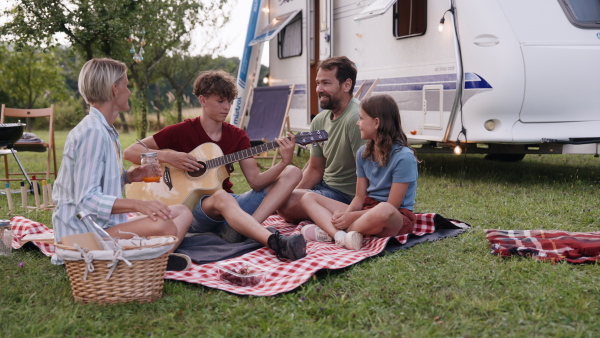 Son playing for family members on guitar. Family traveling in a caravan, camping and having picnic, sitting on blanket.