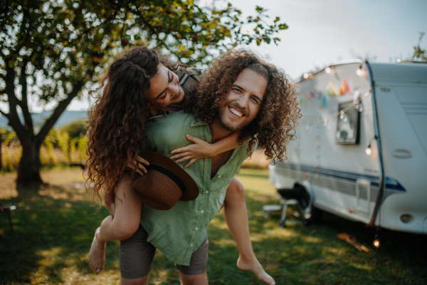 Man in love piggibacking his girlfriend. Young couple is on a camping trip in nature traveling by caravan