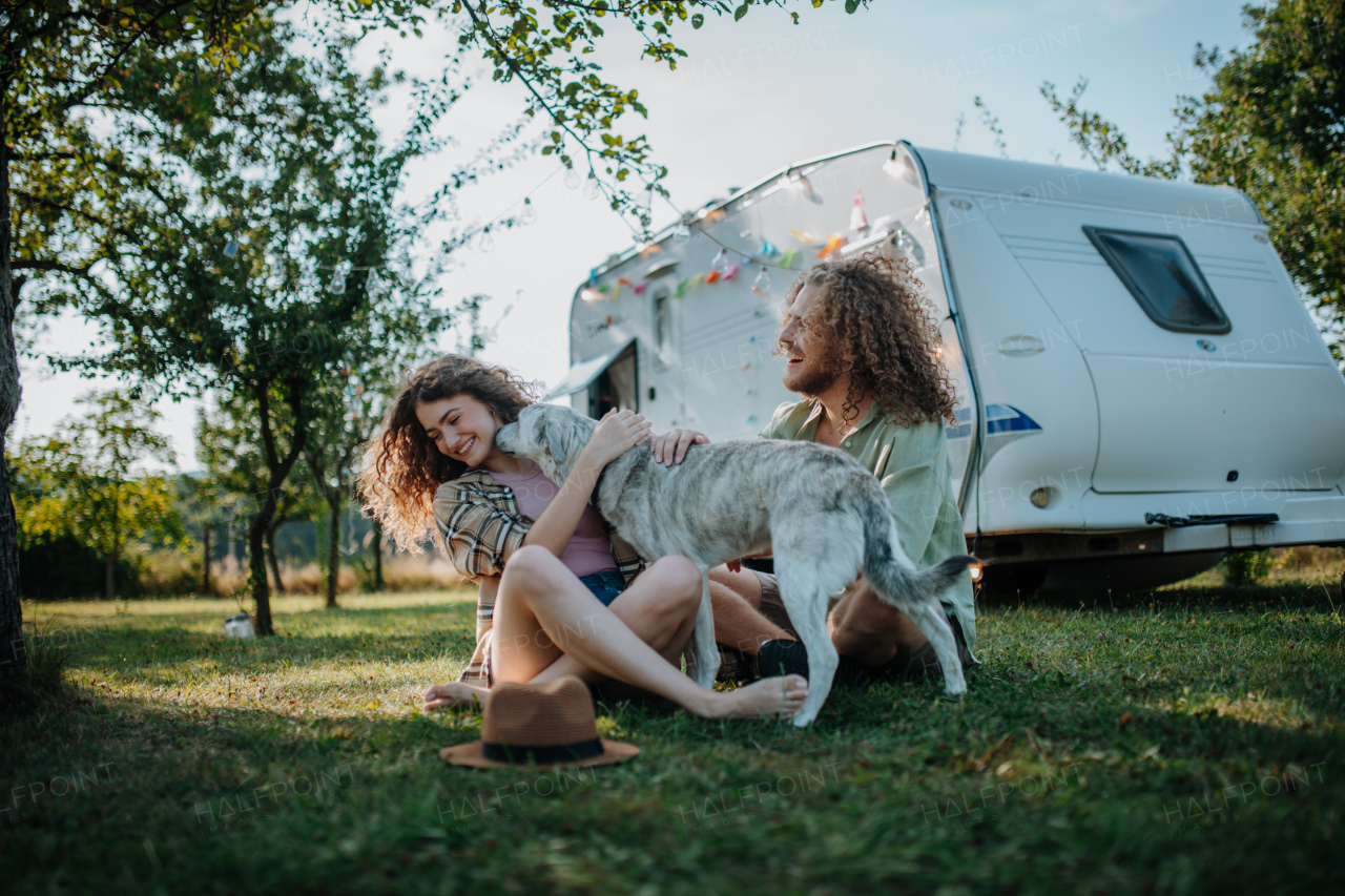 Young couple is on a camping trip in nature playing with dog, sitting in front of camper. Caravan traveling for young people