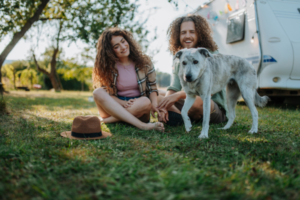 Young couple is on a camping trip in nature playing with dog, sitting in front of camper. Caravan traveling for young people