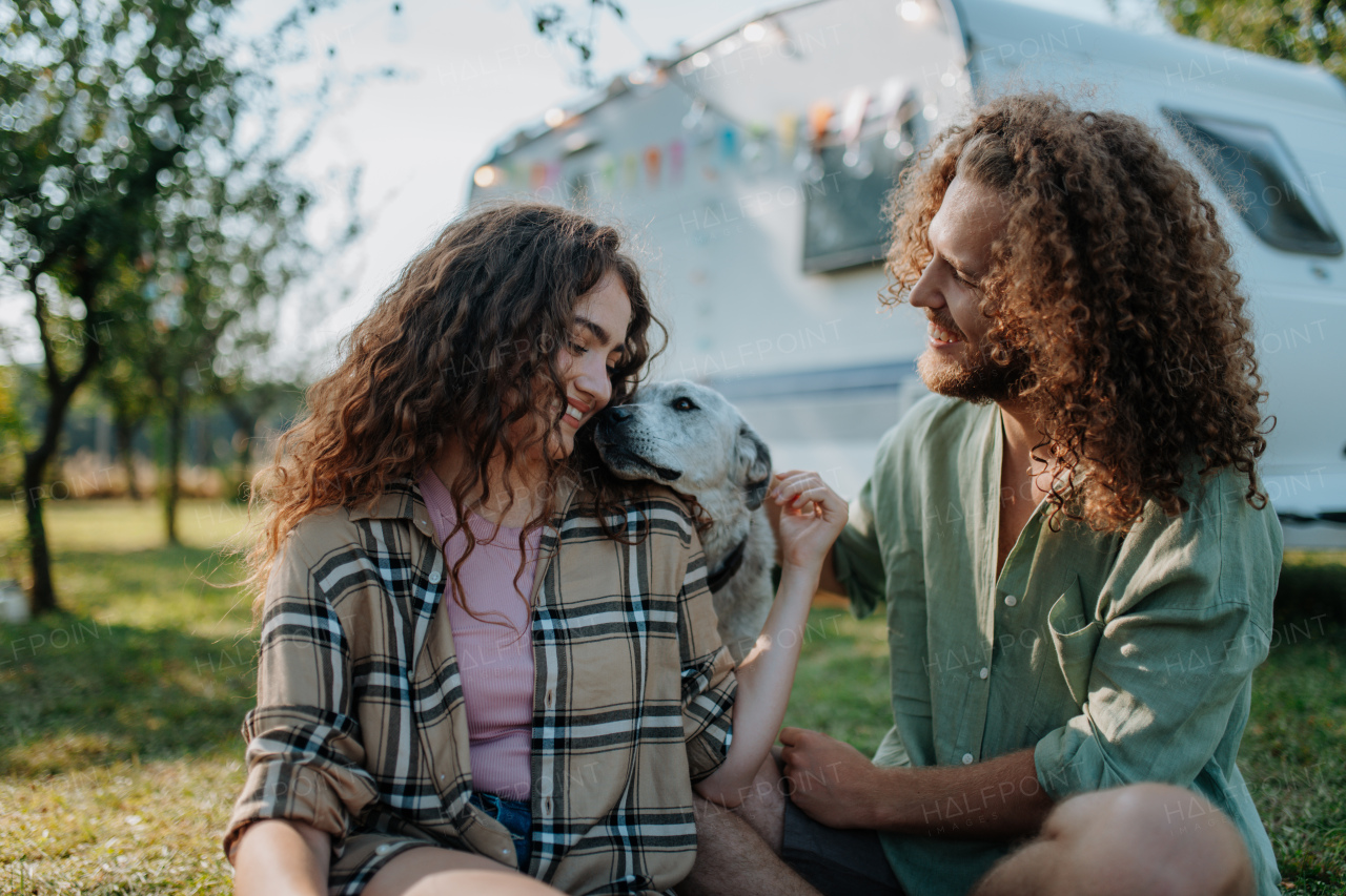 Young couple is on a camping trip in nature playing with dog, sitting in front of camper. Caravan traveling for young people