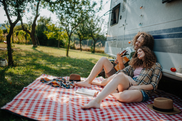 Couple sitting by caravan, enjoying peaceful moment. Man playing guitar and singing song to his girlfriend.