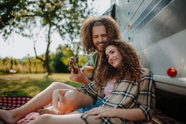 Couple sitting by caravan, enjoying peaceful moment. Man playing guitar and singing song to his girlfriend.