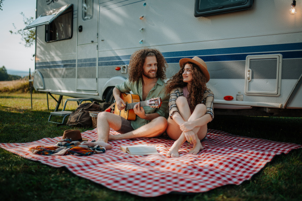 Couple sitting by caravan, enjoying peaceful moment. Man playing guitar and singing song to his girlfriend.