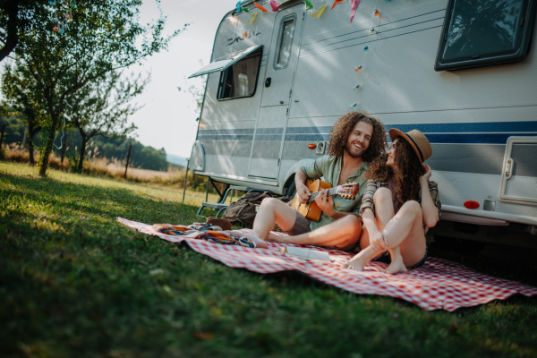 Couple sitting by caravan, enjoying peaceful moment. Man playing guitar and singing song to his girlfriend.