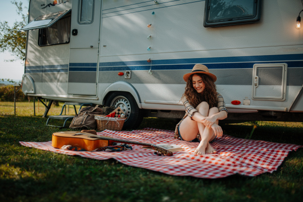 Young beautiful woman is on a camping trip in nature, sitting in front caravan, reading book.