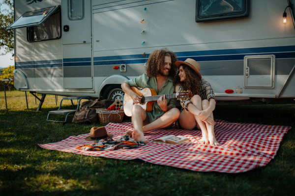 Couple sitting by caravan, enjoying peaceful moment. Man playing guitar and singing song to his girlfriend.