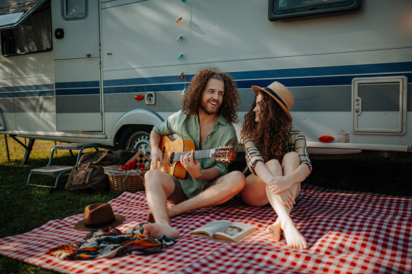 Couple sitting by caravan, enjoying peaceful moment. Man playing guitar and singing song to his girlfriend.