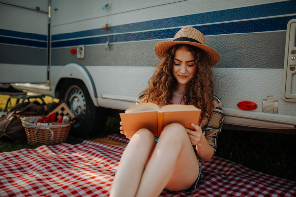 Young beautiful woman is on a camping trip in nature alone, sitting in front caravan, reading book.