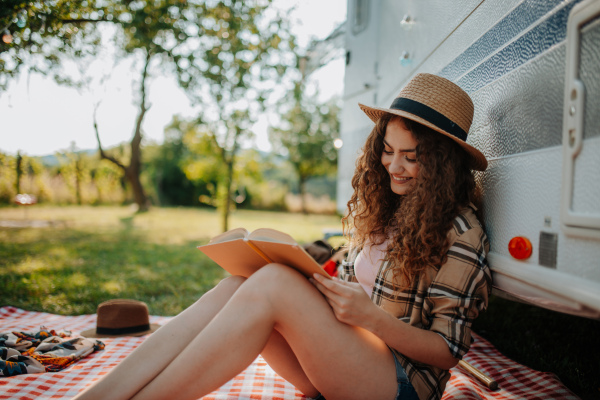 Young beautiful woman is on a camping trip in nature, sitting in front caravan, reading book.