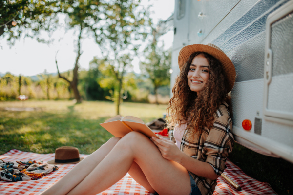 Young beautiful woman is on a camping trip in nature, sitting in front of caravan and reading book. Caravan traveling for young people