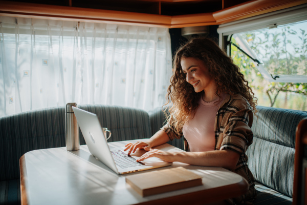 Young beautiful woman is camping in nature, sitting in her caravan and working on notebook. Caravan as homeoffice, working remotely.