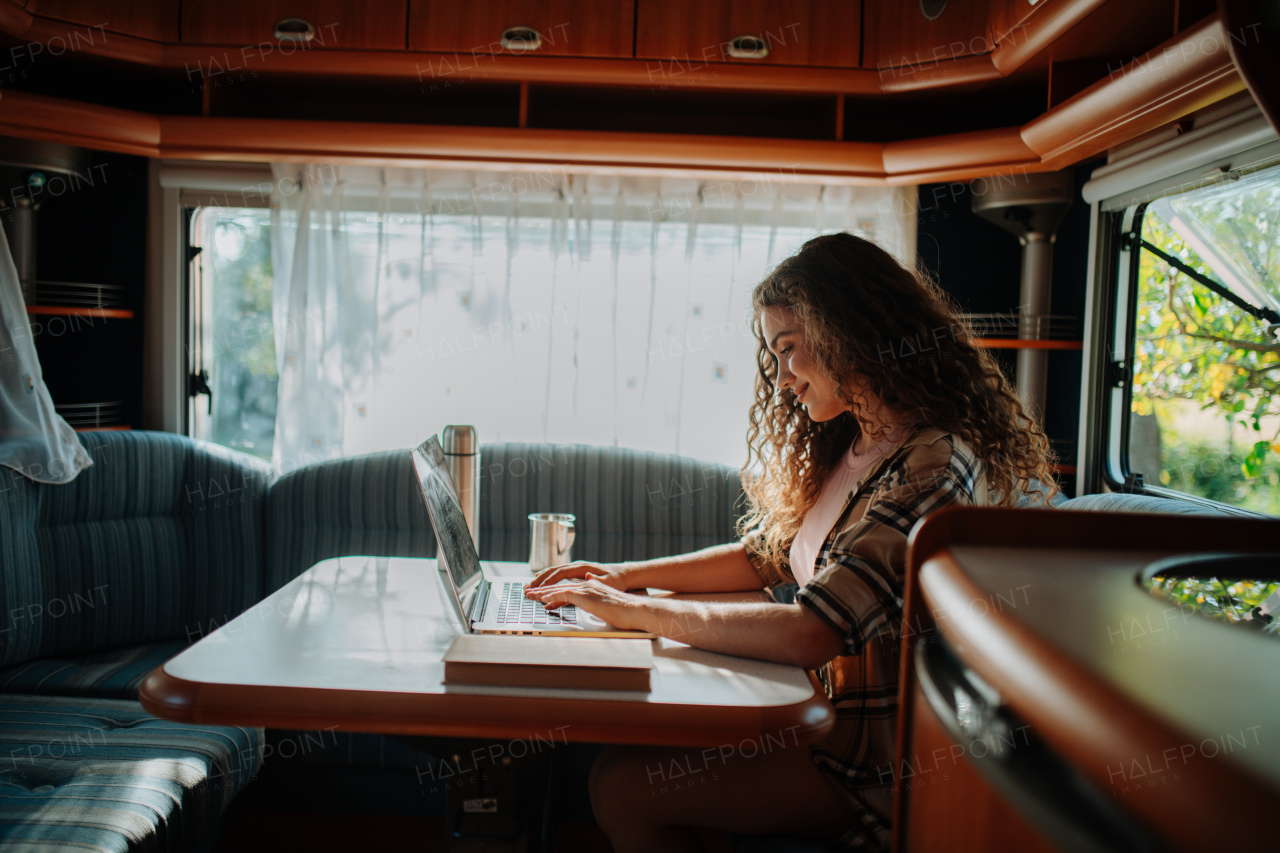 Young beautiful woman is on a camping trip in nature, sitting in her caravan and working on notebook. Caravan as homeoffice, working remotely.