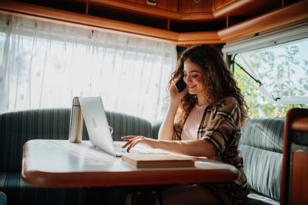 Young beautiful woman is on a camping trip in nature, sitting in her caravan making phone call. Caravan as homeoffice, working remotely