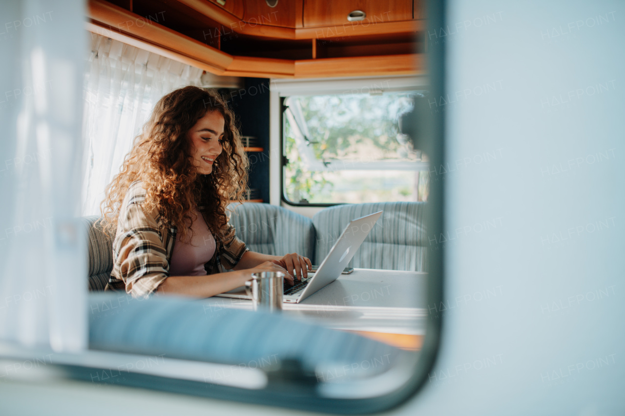 Young beautiful woman is camping in nature, sitting in her caravan and working on notebook. Caravan as homeoffice, working remotely. Through window shot.