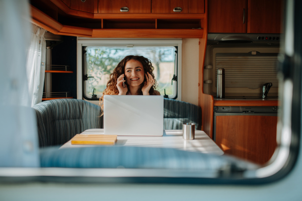 Young woman is camping in nature, sitting in her caravan and working on notebook. Caravan as homeoffice, working remotely.