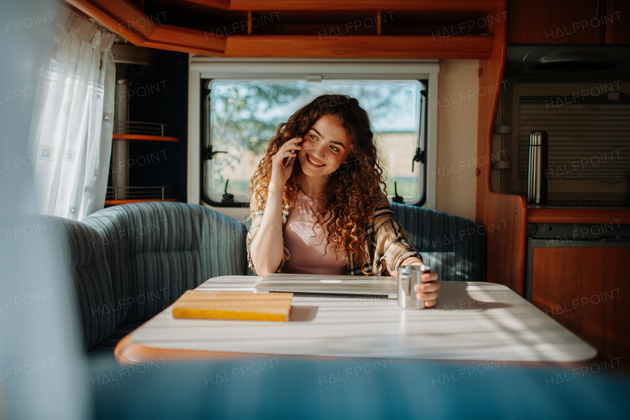 Young beautiful woman is on a camping trip in nature, sitting in her caravan making phone call. Caravan as homeoffice, working remotely