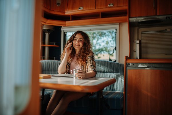 Young beautiful woman is on a camping trip in nature, sitting in her caravan making phone call. Caravan as homeoffice, working remotely