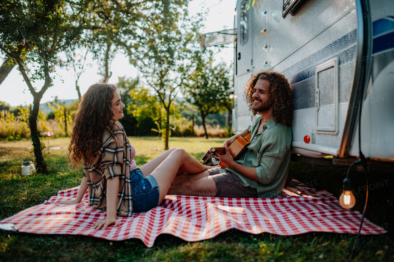 Couple sitting by caravan, enjoying peaceful moment. Man playing guitar and singing song to his girlfriend.