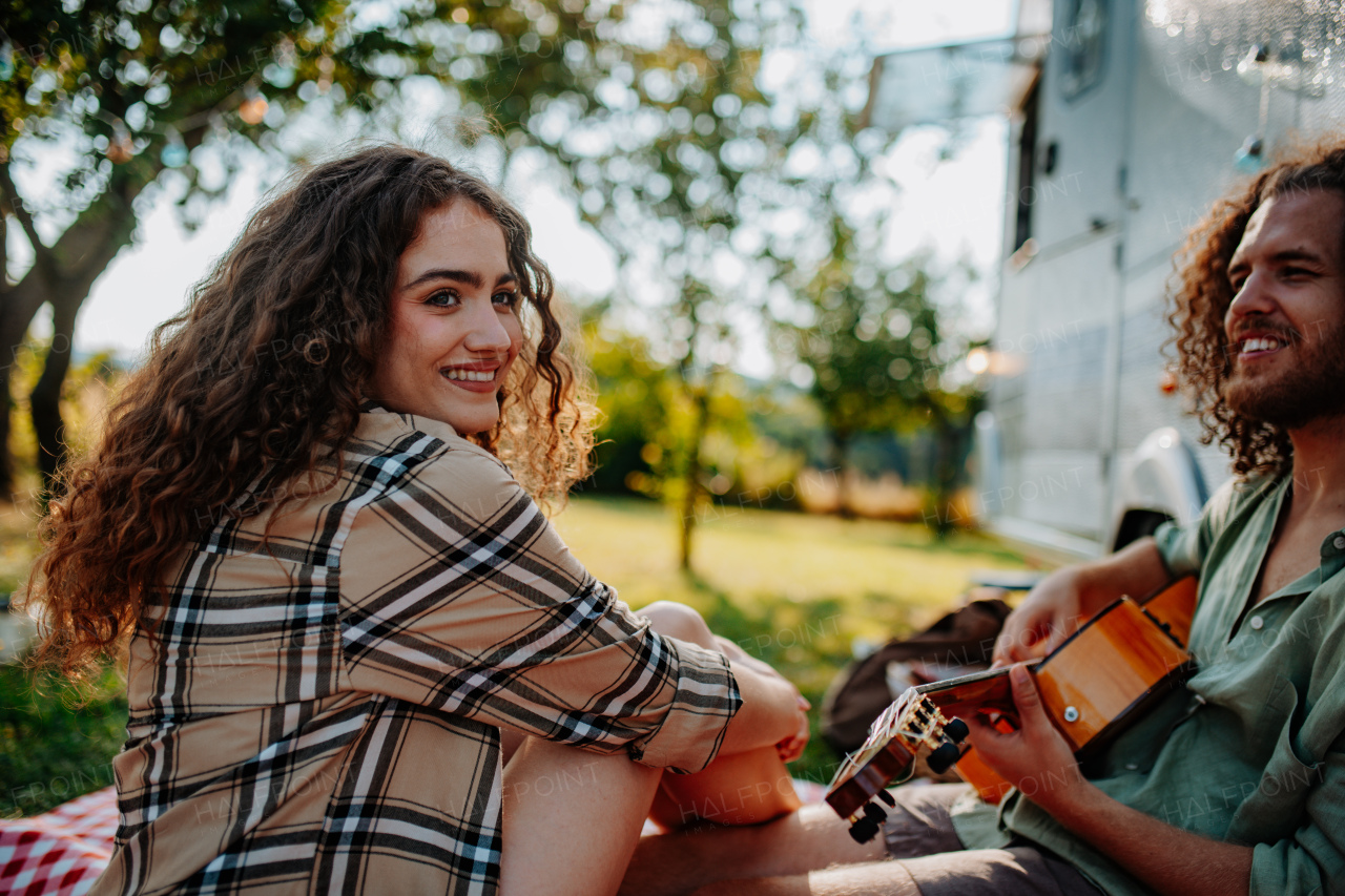 Couple sitting by caravan, enjoying peaceful moment. Man playing guitar and singing song to his girlfriend.