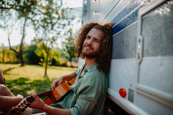 Young man is on a camping trip in nature, sitting in front caravan and playing guitar alone